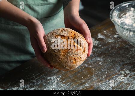 Ein Bild von einer Bäckerin in einer Küche, die einen frisch gebackenen, handwerklichen Brotlaib horizontal in den Händen hält. Stockfoto