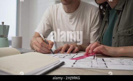 Das junge Ehepaar diskutiert aktiv über Reparaturen in einer neuen Wohnung oder einem neuen Haus, misst die Maße im Diagramm mit einem Lineal, sitzt am Tisch und macht Notizen in einem Notizbuch Stockfoto
