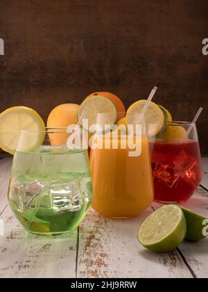 Foto zeigt verschiedene frische Früchte wie Mangos, Zitronen, Limetten und Orangen. In der Mitte des fotos befinden sich 3 Mocktails auf einem weiß getündelten Tisch in einem Glas mit einem W Stockfoto