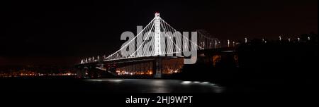 Ein landschaftliches Panorama von San Franciscos Bay Bridge, das weiß leuchtet und nachts mit städtischen Lichtern auf das Meer blickt. Stockfoto