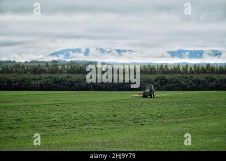 Ein Traktor, der ein Feld mit Bäumen und Bergen im Hintergrund pflügt Stockfoto