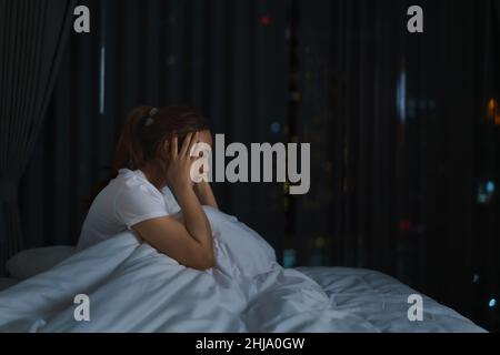 Asiatische Frau, die auf einem weißen Bett im Schlafzimmer liegt und besorgt aussieht oder über etwas über ihr Leben oder ihre Arbeit in der Nacht zu Hause nachdenkt. Stockfoto