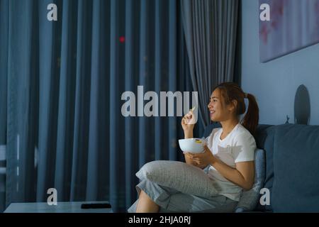 asiatische Frau Fernsehen Spannung Film oder Nachrichten schauen glücklich und lustig und essen Popcorn spät in der Nacht zu Hause Wohnzimmer Couch während der Zeit der Stockfoto