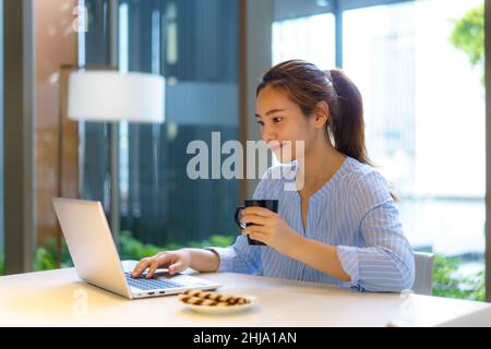 Asiatische Frau trinkt Kaffee in der Tasse und lächelt glücklich, während sie eine Pause von der Arbeit an ihrem Computer während der Arbeit zu Hause im Wohnzimmer nimmt. Stockfoto