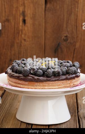 Leckere hausgemachte Schokoladenkuchen mit Ganache, dekoriert mit vielen frischen Brombeeren, Zitronenschale und Puderzucker in weißem Sockel auf Holztisch Copysp Stockfoto