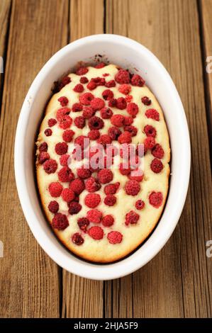 Hausgemachter frisch gebackener Biskuitkuchen mit wilden Himbeeren in weißer ovaler Backform auf altem rustikalem Tisch vertikal Stockfoto