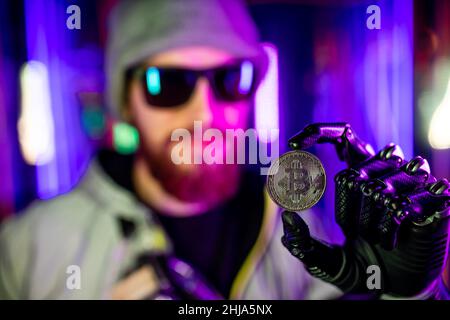 Mann mit prothetischer Roboterhand, die Bitmünze golden im Neon-Hintergrund hält Stockfoto