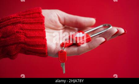Nahaufnahme der Hand einer Frau in einem roten Pullover mit einem Vorhängeschloss mit einem Herz und Schlüsseln auf einem roten Hintergrund. Das Konzept des Valentinstag, Hochzeit, ein Symbol Stockfoto