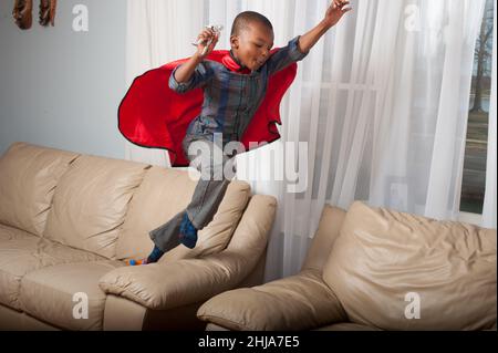 4-jähriger Junge mit Superhelden-Umhang, der von der Couch auf den Stuhl springt und Action-Figur hält Stockfoto