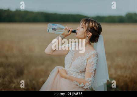 Mädchen im Hochzeitskleid und weißen Schleier trinken Champagner aus der Flasche. Stockfoto