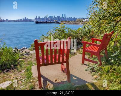 Zwei leuchtend rote Adirondack-Stühle im Hafen von Vancouver Park, Kanada. Zwei rote komfortable Liegestühle am Wasser Stockfoto