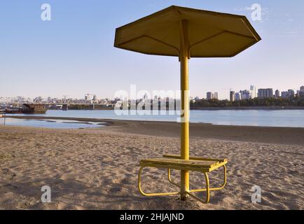 Sonnenschirm-Baldachin am herbstlichen Sandstrand am Ufer des ob in der Hauptstadt Sibiriens. Eine große Stadt mit einer Brücke am Horizont. Blauer Himmel. R Stockfoto