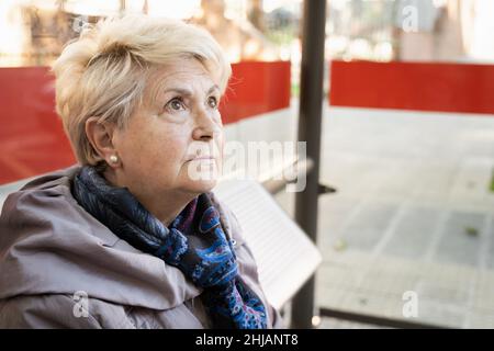Porträt einer älteren blonden Frau, die an einer Bushaltestelle sitzt. Ernsthafte ältere Dame wartet auf öffentliche Verkehrsmittel Stockfoto