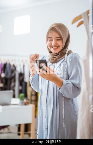 Aufgeregt asiatische muslimische Frau Kleinunternehmen Eigentümer Blick auf Telefon Stockfoto