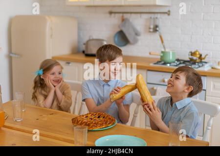 Fröhliche Kinder sitzen an einem Tisch in der Küche, spielen mit Laib Brot, während sie auf Essen warten. Zwei Brüder und Schwester haben Spaß beim Abendessen zu Hause. Familie verbringt am Wochenende Zeit zusammen zu Hause Stockfoto