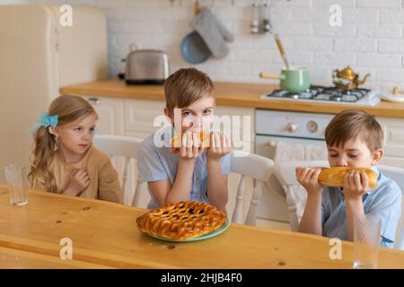 Fröhliche Kinder sitzen an einem Tisch in der Küche, spielen mit Laib Brot, während sie auf Essen warten. Zwei Brüder und Schwester haben Spaß beim Abendessen zu Hause. Familie verbringt am Wochenende Zeit zusammen zu Hause Stockfoto