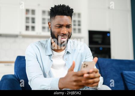 Fokussierter bärtiger multirassischer Mann, der zu Hause auf dem Sofa sitzt und sein Smartphone hält. Kerl, der Nachrichten online liest, Freizeit in sozialen Netzwerken verbringt, dunkelhaarige männliche SMS schreibt und online chattet Stockfoto