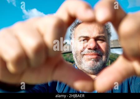 Nahaufnahme eines glücklichen, aufrichtigen bärtigen Mannes mittleren Alters, der mit den Fingern eine Herzgeste macht, Liebe zeigt und die Kamera anschaut. Stockfoto