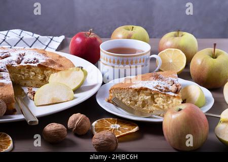 Nahaufnahme einer großen Portion Apfelkuchen mit Walnüssen, umgeben von reifen Äpfeln. Ein Stück Kuchen auf dem Teller und Tee mit Zitrone. Hausgemachte Kuchen mit Saison Stockfoto