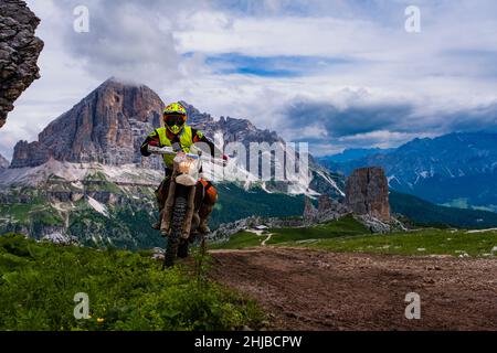 Ein Mann, der mit einem Cross-Motorrad auf die Berghütte Refugio Averau, das Rifugio Scoiattoli und Cinque Torri unten fährt, Tofana-Gruppe in der Ferne. Stockfoto