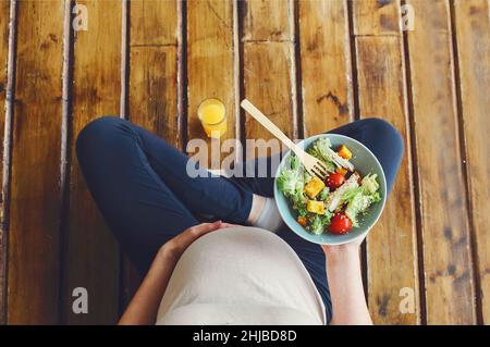 Blick von oben auf die Schwangere mit frischer Salatschüssel und Orangensaft, die werdende Mutter mit gesundem Mittagessen, während sie im Lotus auf dem Holzboden sitzt Stockfoto