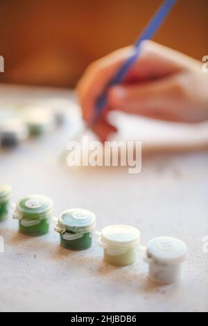 Verschwommenes beschnittenes Foto von weiblicher Handmalerei mit Pinsel auf Papier, selektiver Fokus auf Acrylfarben in Kunststoffkisten, die nach Shad in Reihe angeordnet sind Stockfoto