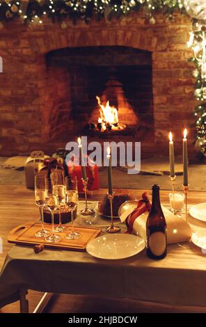 Weihnachts- oder Neujahrsabendessen gegen brennenden Kamin und Weihnachtsgeschenke im gemütlichen Haus mit LED-Lichtern, Festtisch mit frisch gebackenem Kuchen, Stockfoto