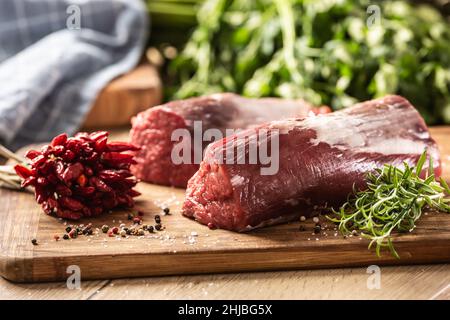 Rohe Stücke von rotem Sirloin-Fleisch auf einem Holzbrett neben frischem Rosmarin und Chilischoten. Stockfoto