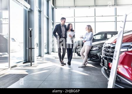 Das junge Paar steht im Autohaus und diskutiert Optionen mit einer Verkäuferin, die sich gegen eines der Autos lehnt. Stockfoto