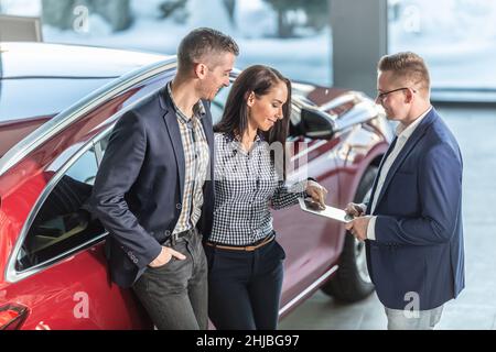 Eine Frau aus einem Paar, das sich für den Kauf eines Autos entschieden hat, zeigt einem Autohändler Details auf einem Tablet. Stockfoto