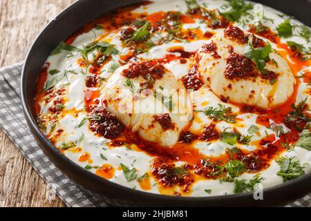 Türkische Eier oder Cilbir werden mit griechischem Joghurt kombiniert mit frischen Kräutern und Knoblauchzehen auf dem Teller auf dem Holztisch hergestellt. Horizontal Stockfoto