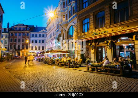 Riga, Lettland. Menschen Ruhen Im Street Cafe Restaurant In Der Altstadt Unter Fassaden Alter Architektonischer Gebäude. Abend- Oder Nachtbeleuchtung In Old Stockfoto