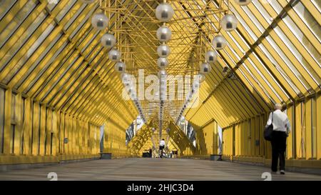 Menschen, die in der Fußgängerbrücke in Moskau, Russland, spazieren gehen. Blick in die Andrejewskij-Brücke mit leuchtend gelben Metallbalken und hängenden Laternen. Stockfoto