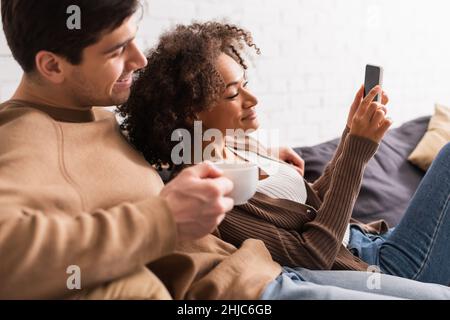 Lächelndes interrassisches Paar mit Kaffee, das zu Hause mit dem Mobiltelefon telefoniert Stockfoto