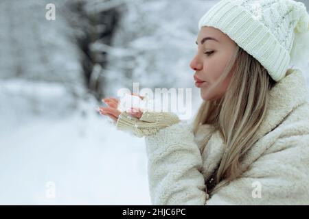 Schönes Mädchen in modischen Winterkleidung bläst Schnee von den Handflächen ihrer Hände. Freude an den ersten Schnee. Junge Blondine in einem weißen Hut in einem verschneiten Stockfoto