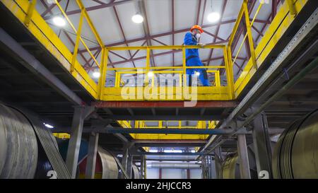 Arbeiter überwacht den Betrieb der Ausrüstung im Werk. Archivmaterial. Ansicht von unten, dass Arbeiter über funktionierende Windenergieanlagen hindurchgehen. Arbeiter überwacht rotierende Tanks. Stockfoto