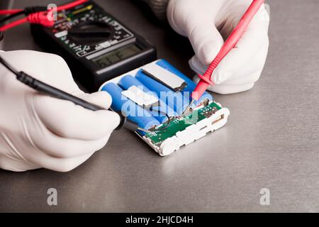 Ladeanzeigeschaltung, rote und schwarze Multimeter-Pole in den Händen von Menschen, die Spannung und Ampere prüfen, 18650 blaue Batterien, grauer Hintergrund. Powerba Stockfoto