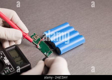 Ladeanzeigeschaltung, rote und schwarze Multimeter-Pole in den Händen von Menschen, die Spannung und Ampere prüfen, 18650 blaue Batterien, grauer Hintergrund. Powerba Stockfoto