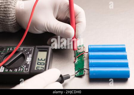 Ladeanzeigeschaltung, rote und schwarze Multimeter-Pole in den Händen von Menschen, die Spannung und Ampere prüfen, 18650 blaue Batterien, grauer Hintergrund. Powerba Stockfoto