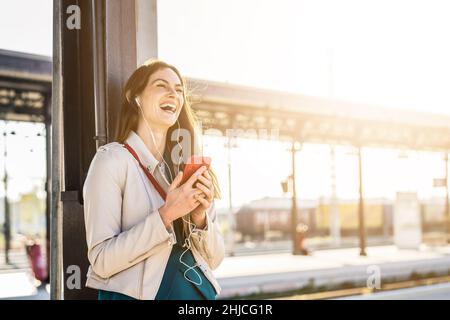 Frau in eleganten Kleidern wartet auf dem Bahnhofssteig mit Smartphone, um die Zeit zu verstreichen. Reise- und Geschäftsfrau-Konzept Stockfoto