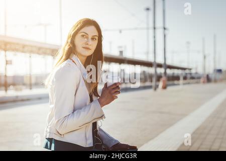 Eine schöne junge Frau im Bahnhof lächelt und wartet auf den Zug am Bahnsteig, sitzt auf dem Gepäck und benutzt das Smartphone Stockfoto