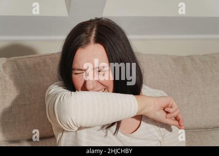 Frau hustet oder niest in ihrem Ellbogen. Ältere Frau mittleren Alters, die zu Hause an allergischer Rhinitis leidet. Symptome von Erkältung oder Allergie. Konzept von sto Stockfoto