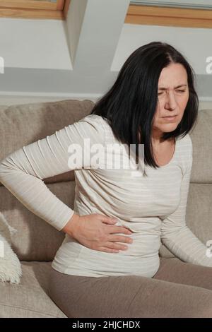 Ältere Frau mittleren Alters, die zu Hause an Rückenschmerzen und Flankenschmerzen leidet. Krankheit, Unmut, Traurigkeit und Unruhe. Symptome von Krankheit oder ungesund lebhaftesten Stockfoto