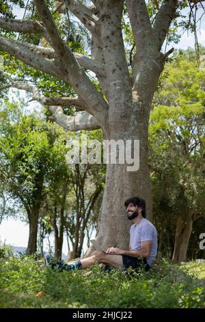 Junger Mann an einem schönen sonnigen Tag in einem Park mit Laptop auf dem Schoß. Graues Notebook für Arbeit und Freizeit. Home Office-Konzept. Grün und Natur zurück Stockfoto