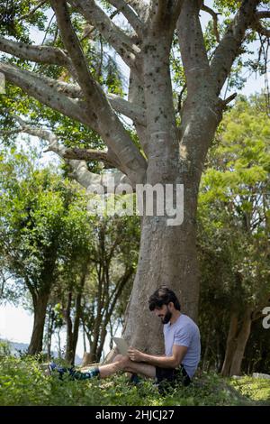 Junger Mann an einem schönen sonnigen Tag in einem Park mit Laptop auf dem Schoß. Graues Notebook für Arbeit und Freizeit. Home Office-Konzept. Grün und Natur zurück Stockfoto