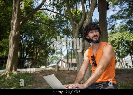 Junger Mann an einem schönen sonnigen Tag in einem Park mit Laptop auf dem Schoß. Graues Notebook für Arbeit und Freizeit. Home Office-Konzept. Grün und Natur zurück Stockfoto