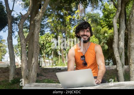 Junger Mann an einem schönen sonnigen Tag in einem Park mit Laptop auf dem Schoß. Graues Notebook für Arbeit und Freizeit. Home Office-Konzept. Grün und Natur zurück Stockfoto