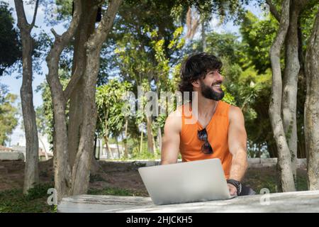 Junger Mann an einem schönen sonnigen Tag in einem Park mit Laptop auf dem Schoß. Graues Notebook für Arbeit und Freizeit. Home Office-Konzept. Grün und Natur zurück Stockfoto