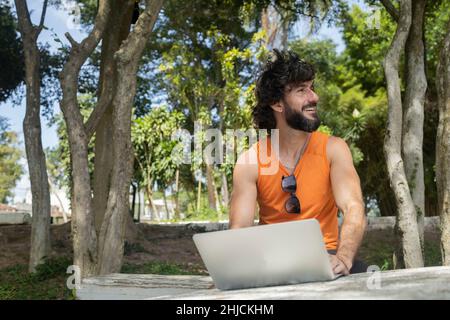 Junger Mann an einem schönen sonnigen Tag in einem Park mit Laptop auf dem Schoß. Graues Notebook für Arbeit und Freizeit. Home Office-Konzept. Grün und Natur zurück Stockfoto