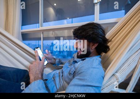 Seitenansicht eines jungen Mannes, der nachts ein Smartphone in seinem Wohnzimmer auf einer Hängematte mit einer Landschaft mit Blick auf die Stadt im Hintergrund verwendet. Mobiltelefon, Technik Stockfoto